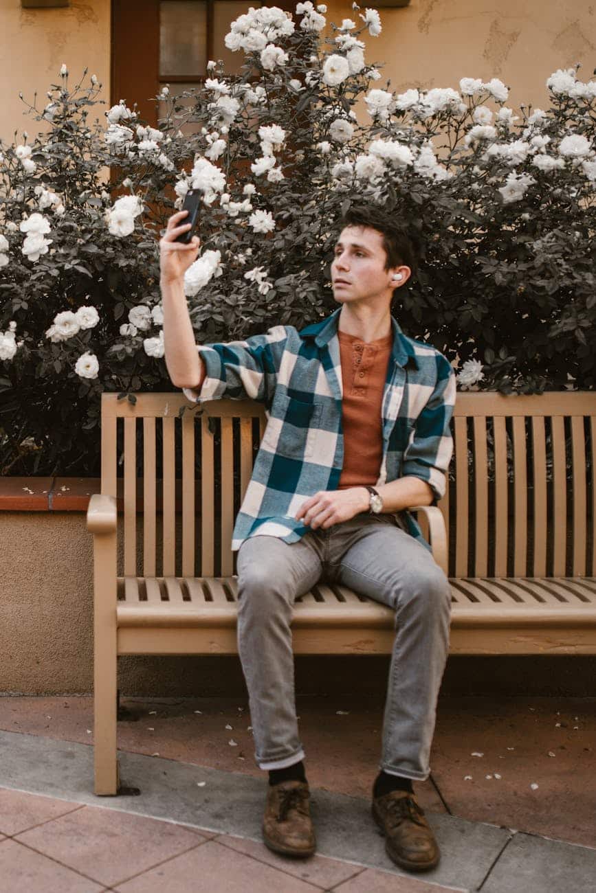 man in blue and red plaid dress shirt sitting on brown wooden bench taking selfie
